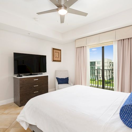 A bright hotel-style bedroom with a large bed, TV on a dresser, a small chair, and sliding doors to a balcony.