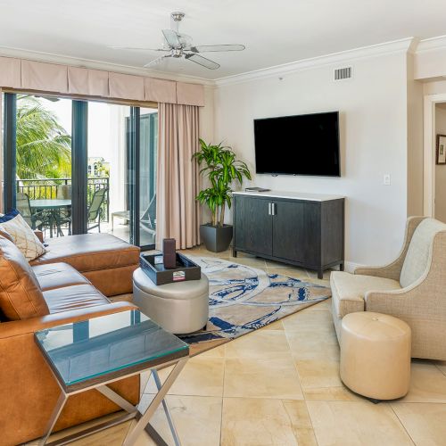 A bright, modern living room with tan leather sofa, a gray TV stand, wall-mounted TV, armchairs, a glass coffee table, indoor plant, and sliding doors to a balcony.
