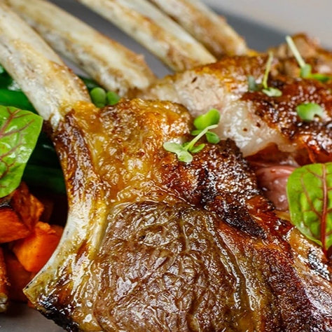 A plate with a roasted lamb rack, chunky carrots, green beans, and pecans on the side, garnished with microgreens and herbs.