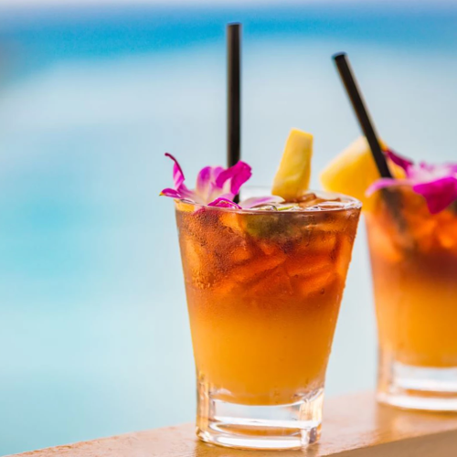 Two tropical cocktails on a ledge by the sea, garnished with flowers and pineapple, with black straws against a blurred blue backdrop.