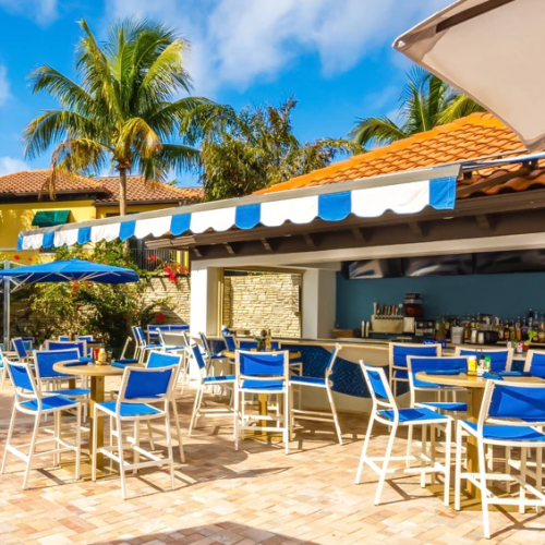 A sunny poolside bar and patio with blue chairs, umbrellas, palm trees, and a thatched roof setting.
