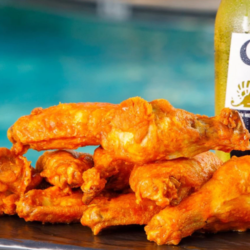 A plate of spicy orange chicken wings with a Corona Extra beer bottle in the background by a pool, tasty bites ready to devour.