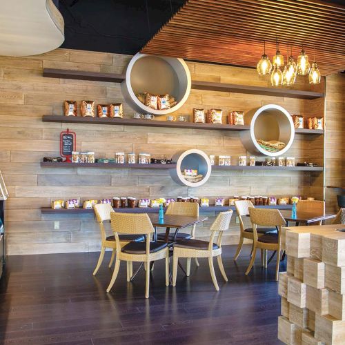 A cozy cafe with wooden walls, round wall shelves, and circular light fixtures; wooden tables and chairs, a counter with pastries, and warm ambient lighting.