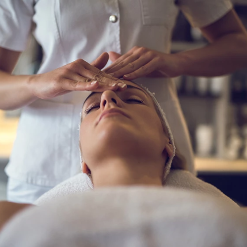 A spa treatment where a practitioner applies a facial or forehead massage on a relaxed client, with head wrapped and eyes closed.