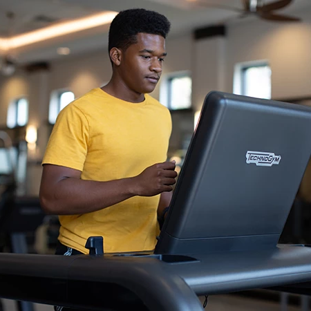 A man in a yellow shirt uses a treadmill desk with a laptop mounted on it inside a gym, focused on the screen while walking.