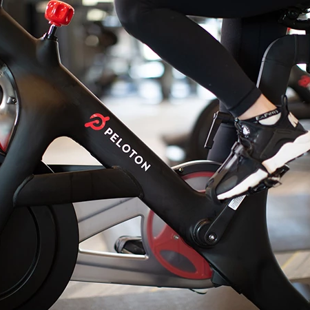 A close-up of a Peloton stationary bike with a rider&rsquo;s foot on the pedal, showing the frame logo and a red water bottle holder, indoors.