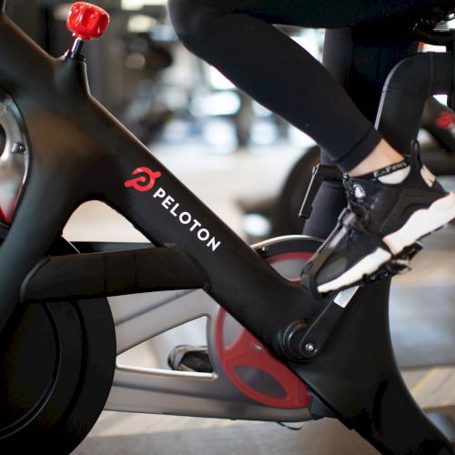 A person is riding a Peloton indoor bike trainer, with feet clipped into pedals and sneakers visible on a matte black frame.
