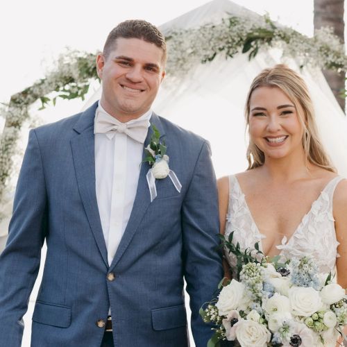A newlywed couple stands smiling in wedding attire&mdash;groom in blue suit and bow tie, bride in a white gown holding a bouquet, outdoors.