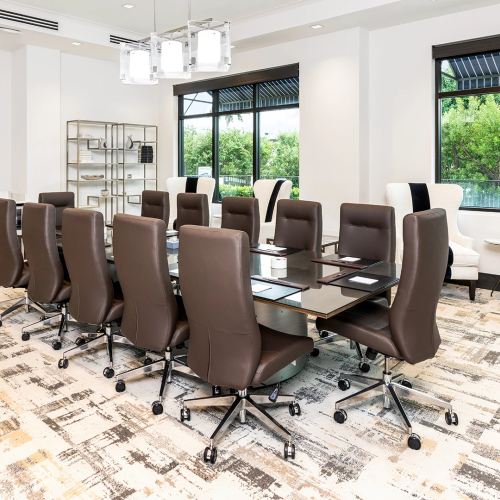A modern conference room with a long dark table, brown chairs, and large windows, plus a seating area and wall decor.