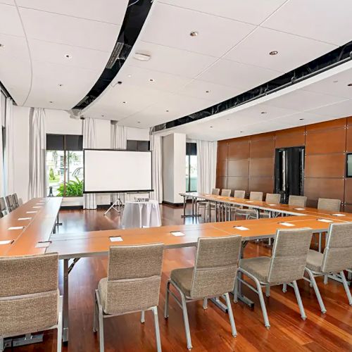 A modern conference room with a U-shaped wooden table, beige chairs, a projector screen, and large windows, perfect for meetings.