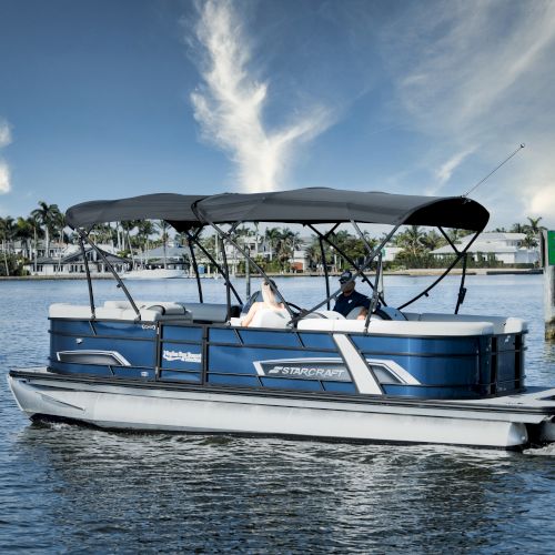 A blue pontoon boat with a canopy glides on calm water near a marina with palm trees and clear skies.