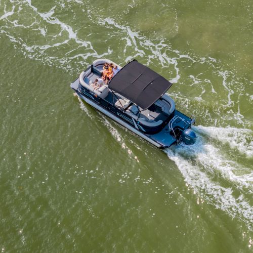 A blue motorboat with a black canopy sails through greenish water, leaving white wake as people ride along in a sunny, open-water scene.
