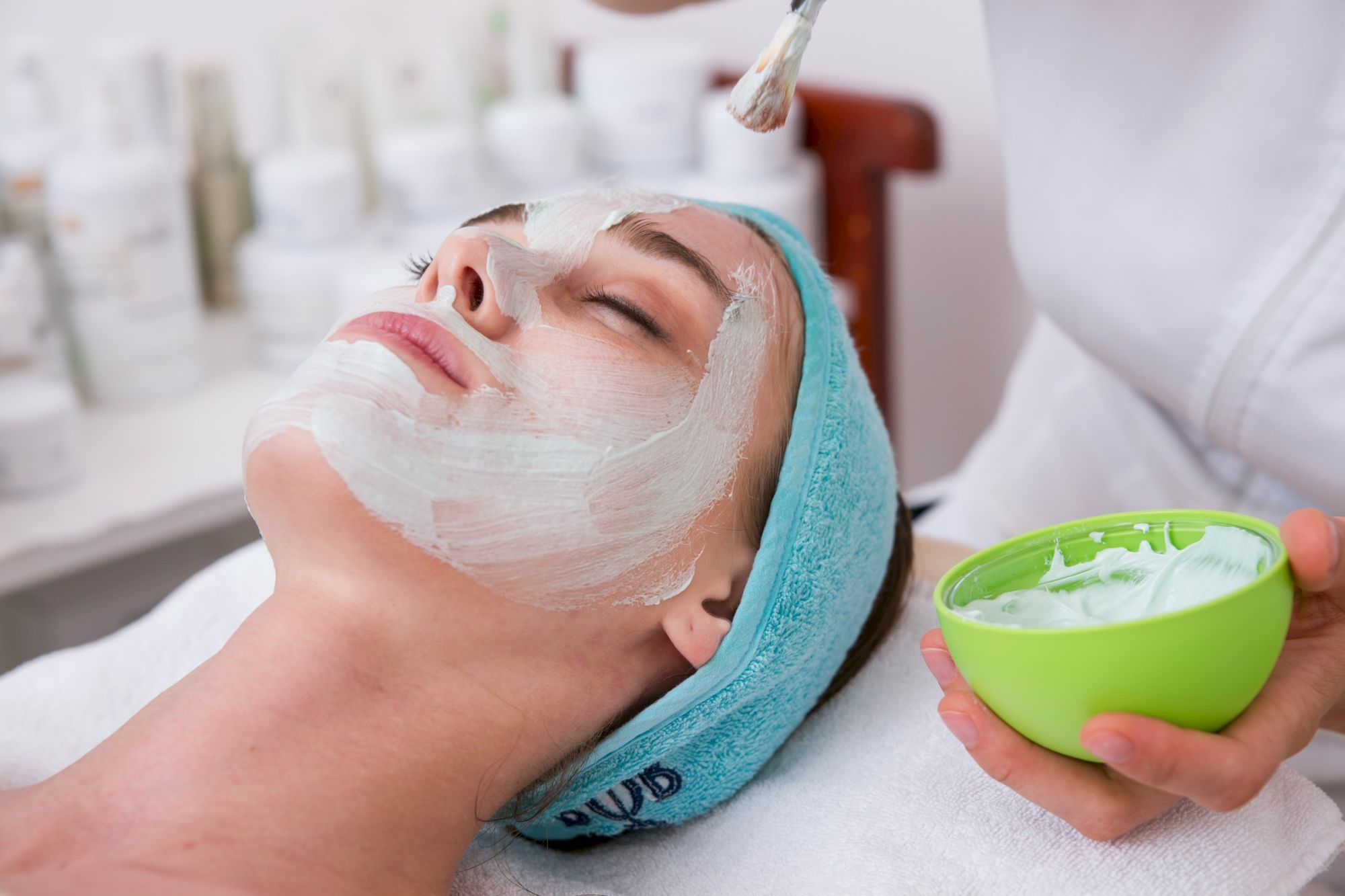 A woman with a spa facial mask relaxing on a treatment bed; a green bowl of mask product sits nearby, with a clinician applying it.