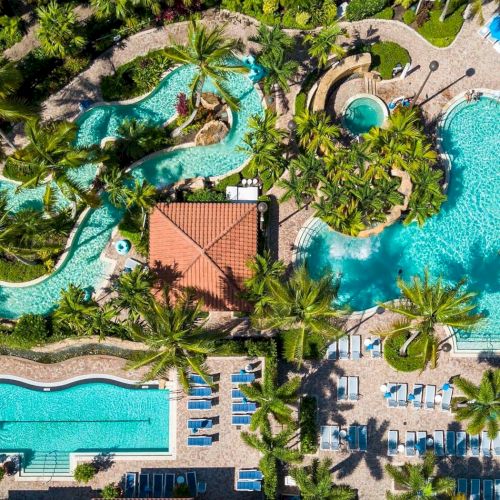 Aerial view of a tropical resort with winding pools, palm trees, sun loungers, and a central building, all surrounding bright blue water.