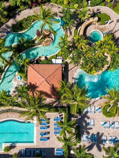 Aerial view of a tropical resort with winding pools, palm trees, sun loungers, and a central building, all surrounding bright blue water.