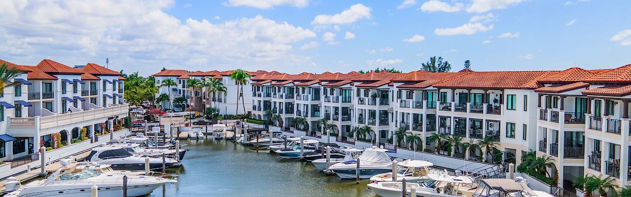 A sunny marina with calm water, lined by white buildings with red-tiled roofs and docked boats on both sides. Ends now.