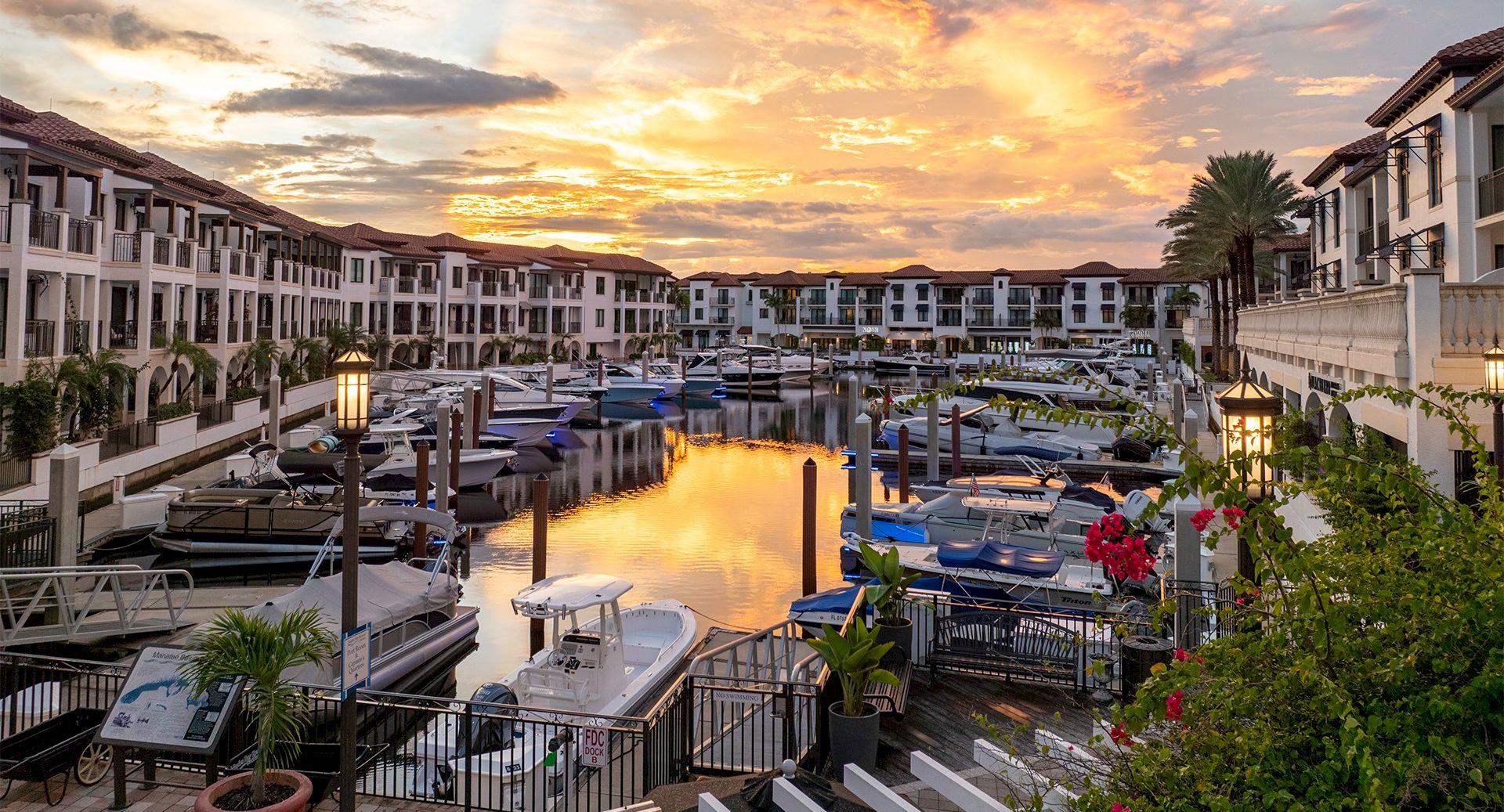 Sunset over a marina with calm water, boats docked, white townhouses, palm trees, and a bridge; warm orange glow reflects off the water.