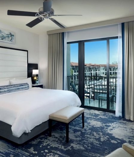 Modern hotel room with a king bed, white headboard, blue-gray rug, ceiling fan, and a balcony view of buildings. End with a period.
