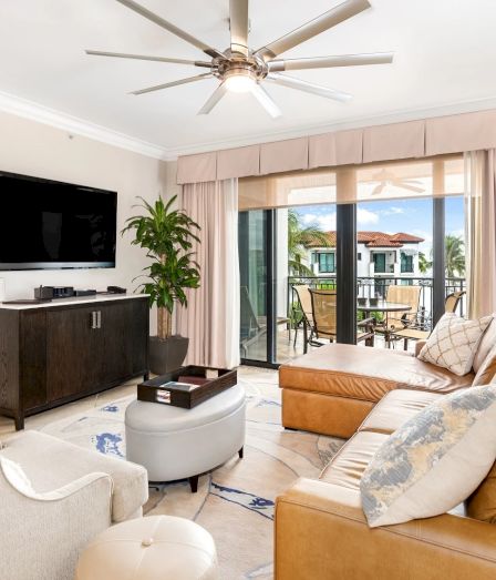 A bright living room with a wall-mounted TV, dark wood media console, cream seating, a tan leather sofa, a glass coffee table, a ceiling fan, and a sliding door to an outdoor patio.