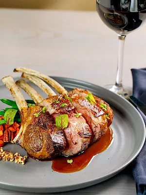 A plated rack of lamb, juicy and seared, with sauce, vegetables, and a glass of red wine in the background.