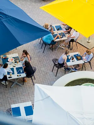 People dine at outdoor cafe tables under colorful umbrellas on a sunny patio.