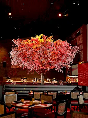 A cozy dining area with a pink blossom tree centerpiece, modern seating, warm lighting, and a large Buddha statue on a red pedestal.