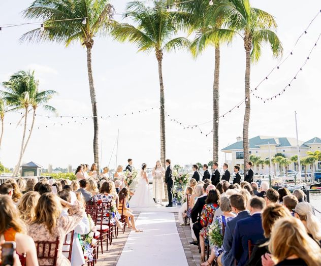 A wedding ceremony outdoors on a pier or aisle with guests seated on either side, palm trees, string lights, and a couple at the altar.