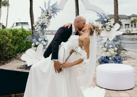 A bride and groom kiss by a lakeside wedding arch, with palm trees, floral decor, and modern seating around a dock.