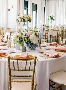 An elegant banquet hall set with round tables, white tablecloths, peach napkins, gold chairs, and lush floral centerpieces for a formal event.
