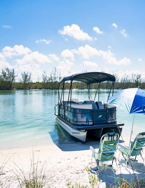 A sunny beach with a small pontoon boat under a canopy, two chairs on the white sand, and a blue beach umbrella by clear turquoise water.