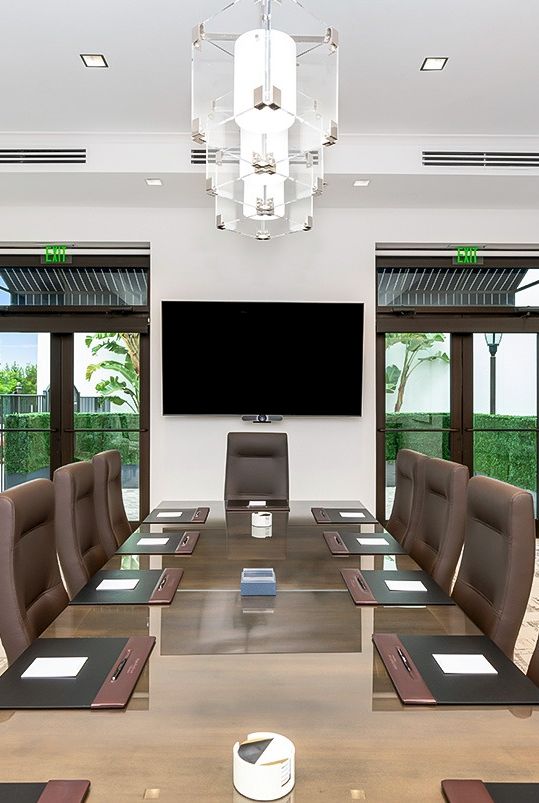 A modern conference room with a long glass table, brown leather chairs, a wall-mounted TV, and large windows opening to greenery. End sentence.