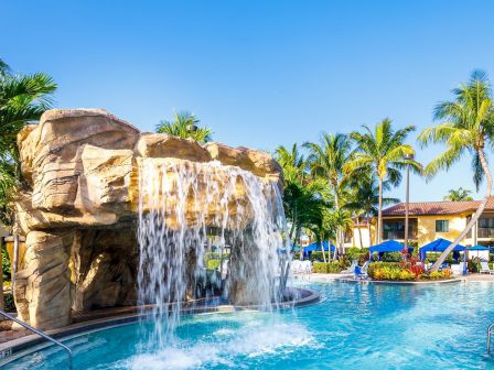 A sunny resort pool with a rocky waterfall, palm trees, and blue umbrellas beside a hotel under a clear blue sky.