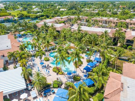 Aerial view of a tropical resort with palm trees and multiple swimming pools, surrounded by sunny houses and gardens.