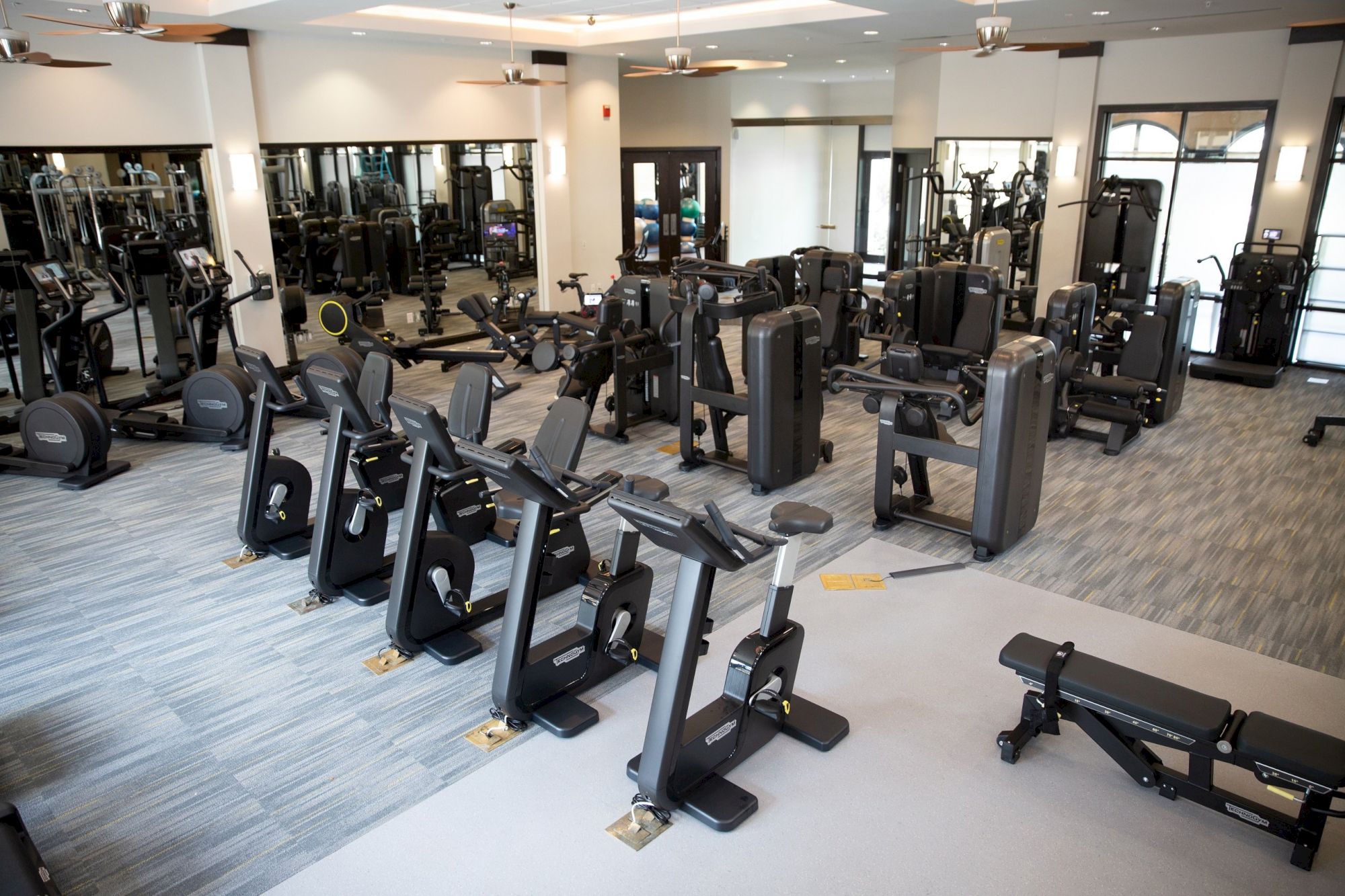 A bright gym full of strength machines and cardio equipment, with ceiling fans and rows of workout stations waiting for use.