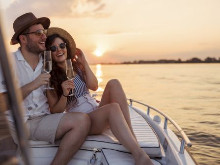 A smiling couple lounges on a boat at sunset, wearing sunglasses and hats, holding champagne flutes by the water.