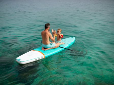 A man paddles a SUP in calm water with a child on board, holding a red-and-yellow life buoy as they splash and wave.