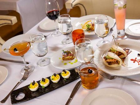 A dining table set with wine glasses and small plated appetizers, plus a tall pink drink in a candle-like holder.