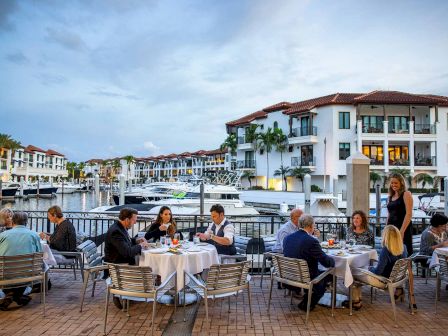 People dine outdoors at a waterfront restaurant with yachts and a white hotel building under a cloudy sky.