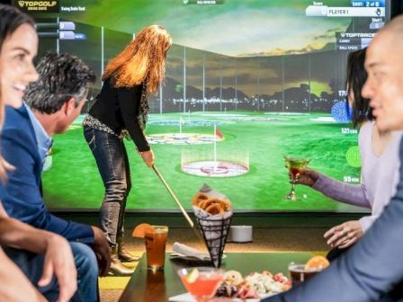 A few people sit at a table watching a golf-simulator screen, with food and drinks on the table.