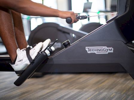 A person runs on a Technogym treadmill, holding the handle with one hand while wearing white sneakers.