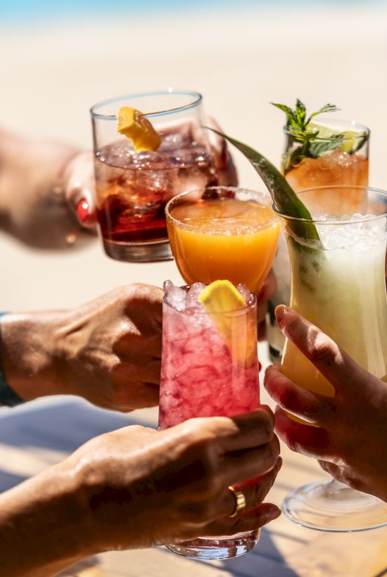 Friends clink glasses of colorful cocktails and juice with garnishes for a sunny toast.