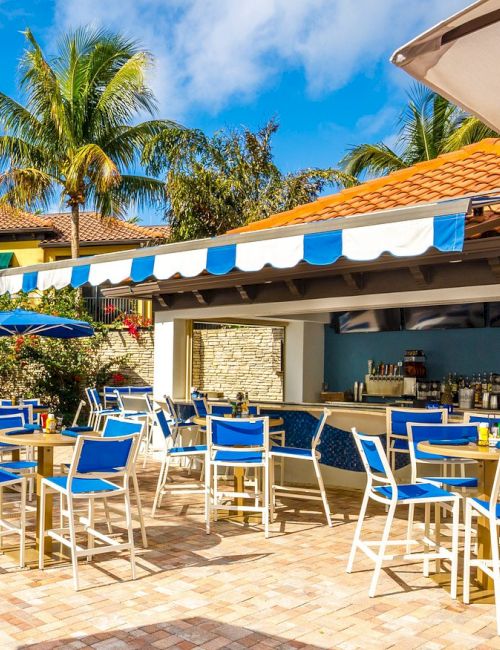 An outdoor hotel patio with blue-and-white chairs, a bar setup, umbrellas, and palm trees under a sunny sky.