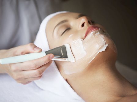 A skincare esthetician applies facial cream with a handheld device while a woman rests with eyes closed.