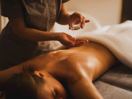 A masseuse pours oil into their hands and massages a client&rsquo;s bare shoulder in a warm spa setting.