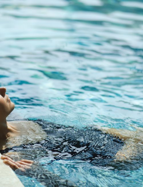 A person lounges in a bright outdoor swimming pool, floating on the edge while sunlight ripples across the water.