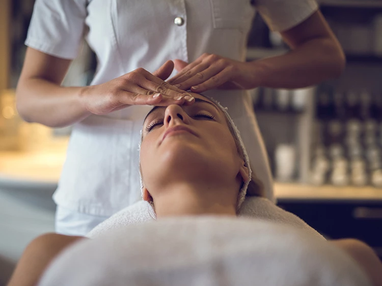 A spa therapist massages a woman&rsquo;s face with hands while she relaxes on a treatment bed.