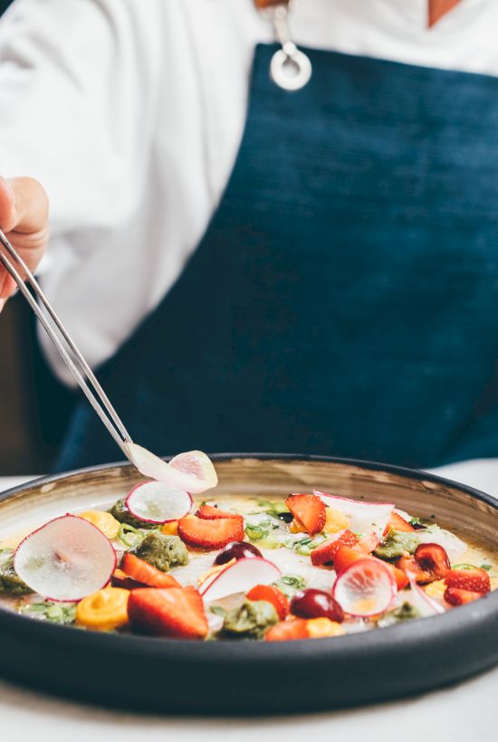 A chef in an apron stirs a bowl of soup with chopsticks, adding radish and diced vegetables.