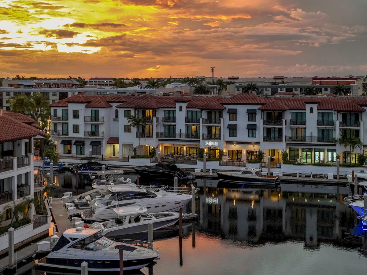 A marina with docked yachts beside a resort building at golden sunset, reflecting in calm water.