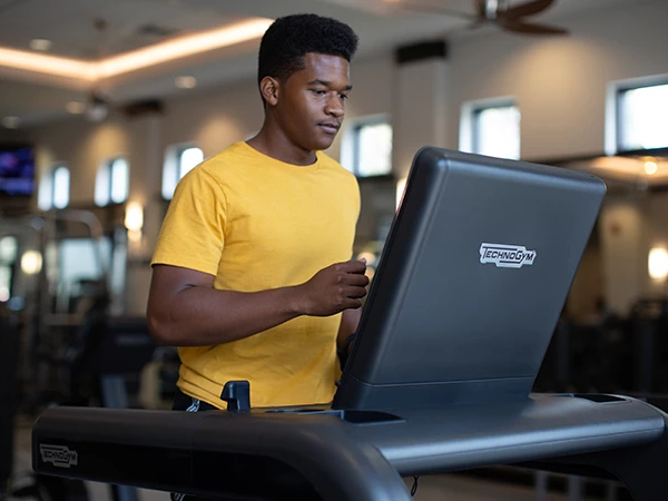 A young man in a yellow shirt uses a treadmill in a gym, standing by the console and focusing ahead.