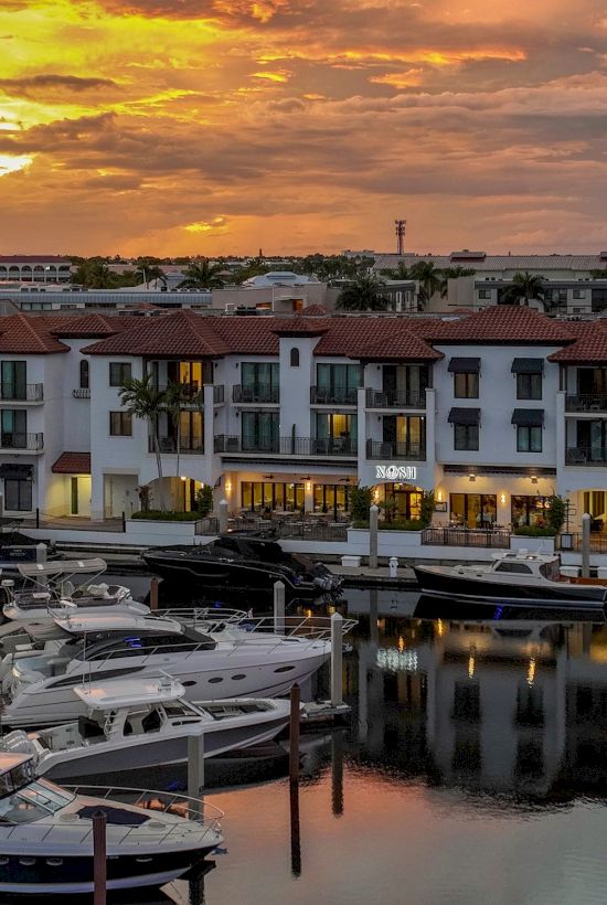 A marina with luxury yachts in the foreground, white hotel buildings behind, and a glowing sunset sky reflecting on calm water.